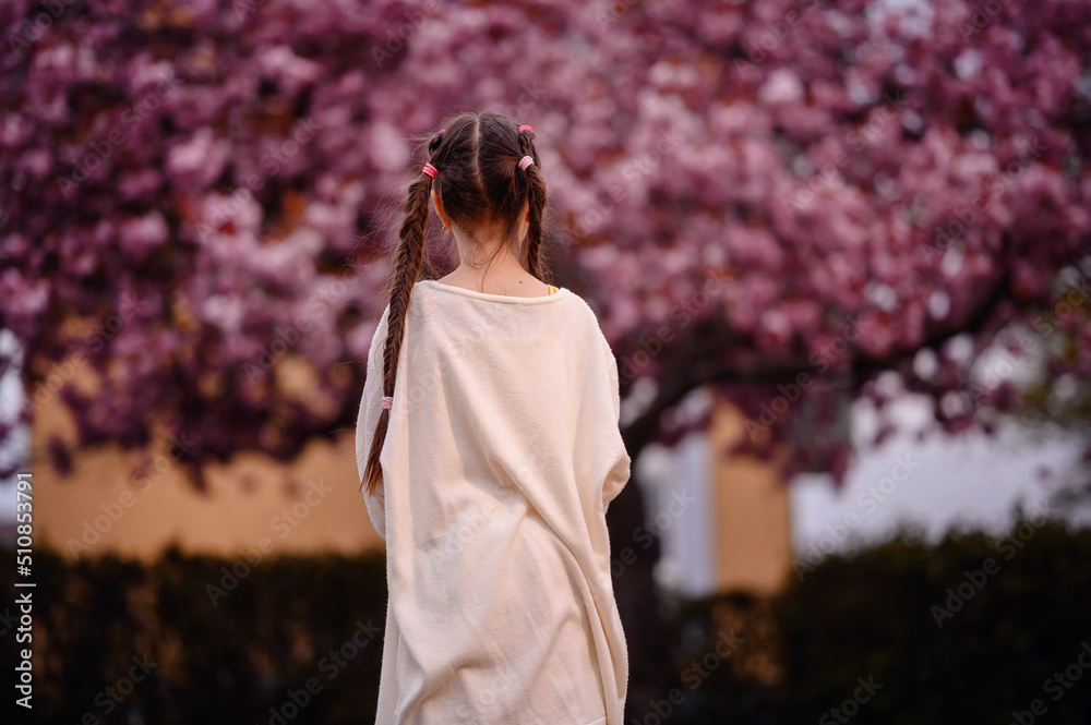 Little preschooler girl in yellow dress, white cardigan with long hair stand before pink sakura tree Little preschooler girl in yellow dress, white cardigan with long hair stand before pink sakura tree