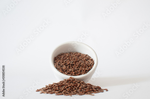 Flaxseeds, profile view of seeds spill from a small, white bowl onto a white surface in a minimalist scene with isolated background.