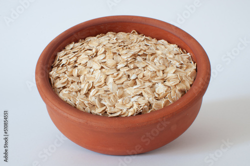 Dried oats, brown earthenware bowl full of lightly-colored oat flakes. Profile view of popular, natural and healthy breakfast meal isolated on clean white background.