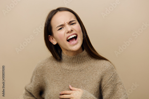 an outraged woman stands on a beige background in a brown sweater  frowns angrily and screams  putting her hand on her chest