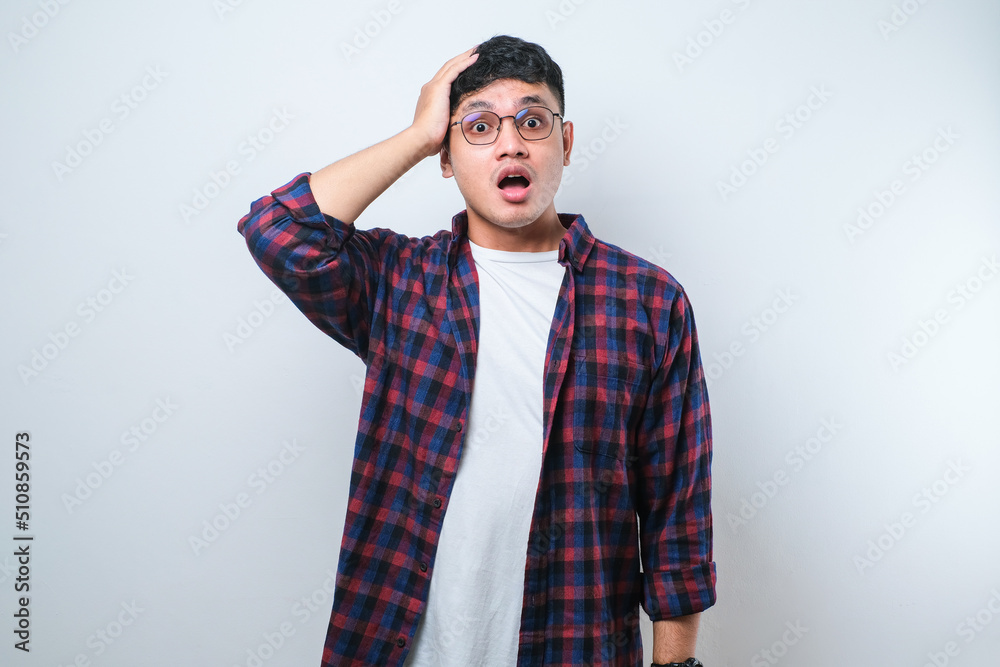 Handsome young Asian man wearing casual shirt and glasses standing with a shocked expression and holding his head