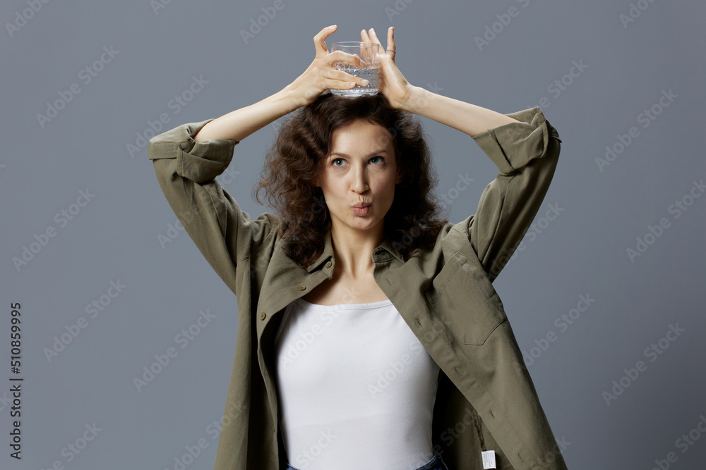 Naklejka premium Funny pretty curly beautiful woman in casual khaki green shirt hold glass of water overhead posing isolated on over gray blue background. Healthy lifestyle. Water is Life concept. Copy space