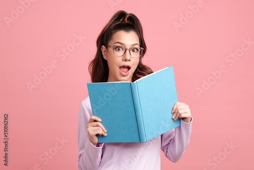 Cute Asian girl is holding the book and standing on pink background with surpirsed face, blank copy space for your advertising content.