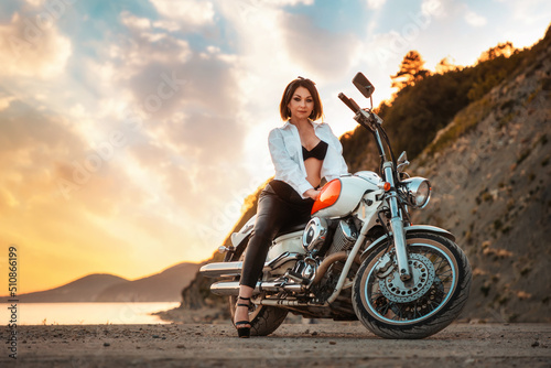 Beautiful sexy adult woman with a high heels and leather pants, posing sitting on motorcycle. Epic sunset sky on the background. The concept of Motorcyclist Day and moto travel
