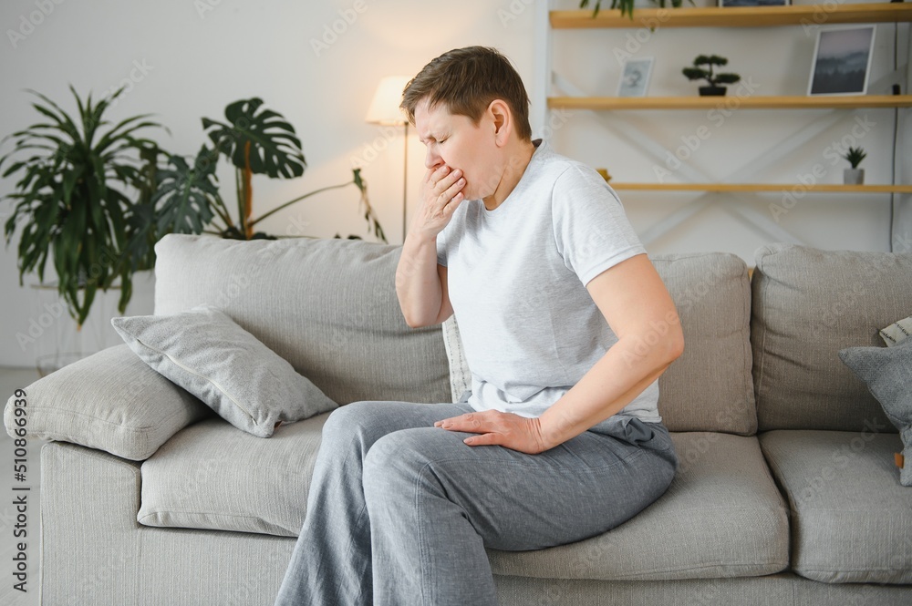 Sick adult woman coughing covering mouth with tissue sitting on a couch at home.