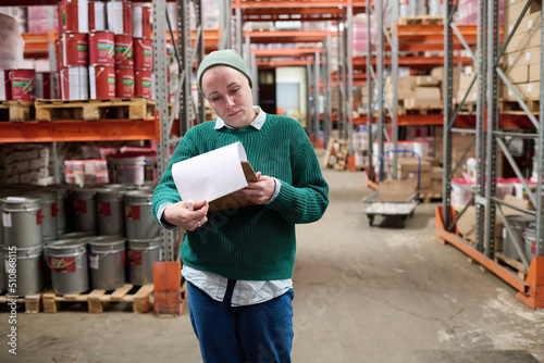 Mature woman examining checklist and controlling the supplies while walking along the warehouse