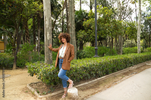 A middle-aged, beautiful, curly-haired Spanish woman stands in a park with large trees in a European city. The woman is dressed in modern and trendy clothes. Travel and holiday concept.