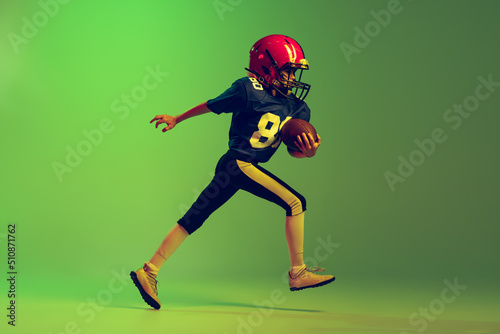 Sportive little boy in sports uniform and equipment playing american football isolated on green background in neon light. Concept of sport, movement, achievements.