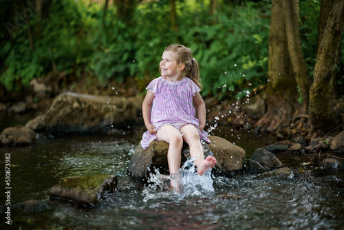 Cute little toddler girl having fun by a river on warm and sunny summer day. Happy excited preschool child splashing with water in forest stream creek.