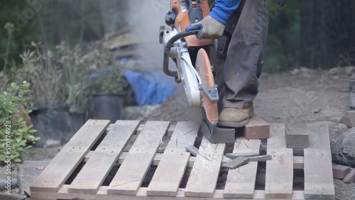 Using a masonry saw, a man holds a red brick paver with his boot as the red dust flies.