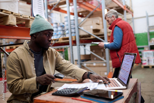 African American distributor using laptop to scan barcodes of parcels while sitting at his workplace at warehouse photo