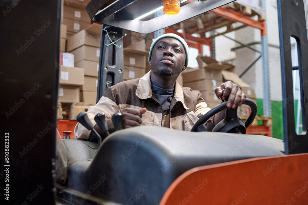 Serious African worker in uniform driving forklift at warehouse to load cargo Serious African worker in uniform driving forklift at warehouse to load cargo