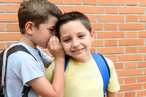 A schoolboy tells something in the ear of another student who laughs about it.