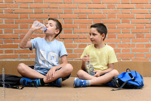 Boys sitting on the ground drinking water from a plastic bottle next to his school bag.