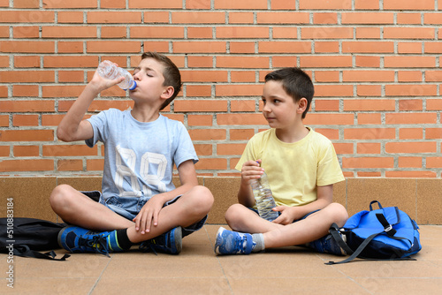 Boys sitting on the ground drinking water from a plastic bottle next to his school bag. photo