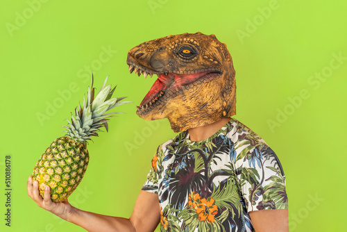 Portrait of happy man wearing dinosaur and hawaiian shirt holding pineapple fruit in hand.Funny celebration with copy space isolated on studio green background.Tropical travel destination 