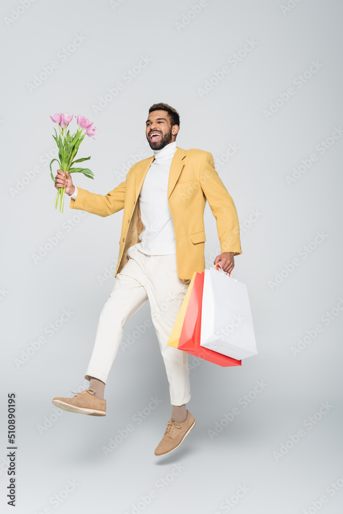 full length of cheerful african american man in yellow blazer and polo neck holding pink tulips and shopping bags on grey. full length of cheerful african american man in yellow blazer and polo neck holding pink tulips and shopping bags on grey.