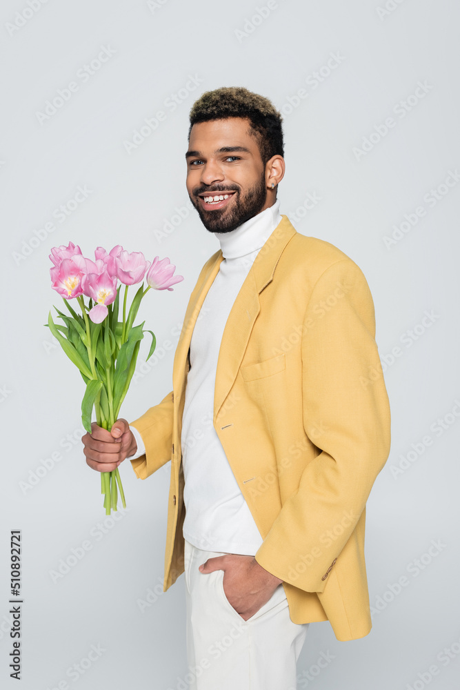 happy african american man in stylish blazer holding bouquet of pink tulips isolated on grey. happy african american man in stylish blazer holding bouquet of pink tulips isolated on grey.