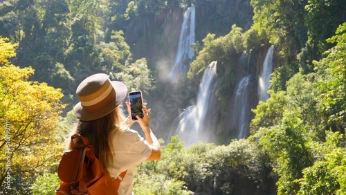 Happy female tourist with backpack in travel. Wanderlust, tourism concept. Young woman traveler make vertical video for social media of amazing waterfalls and tropical nature on her smartphone camera. photo