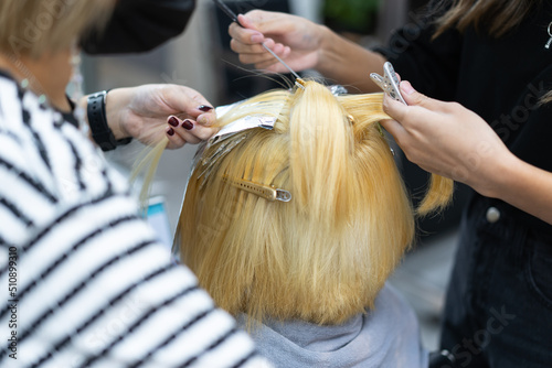 Close-up of two Asian professional female hairdresser's hands using comb dyeing a new color on bleached hair of young short-haired Asian female customer sitting on hairdressing chair in a hair salon.