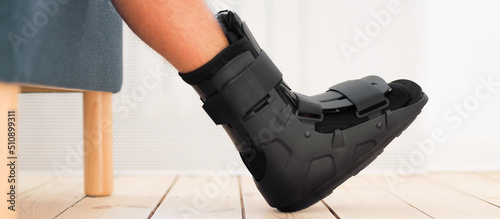 Ankle sprain orthosis, black splint, walker boot on leg sitting on the sofa at home. Ankle foot orthosis close-up.. © mallmo