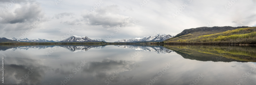 Panoramic view of a wilderness lake in Canada during spring with reflection of snow capped mountains in water. 