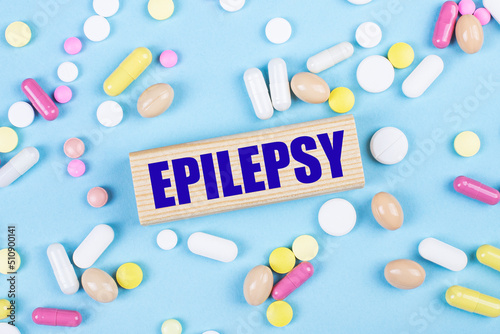 On a blue background, multi-colored pills and a wooden block with the text EPILEPSY. View from above. Medical concept