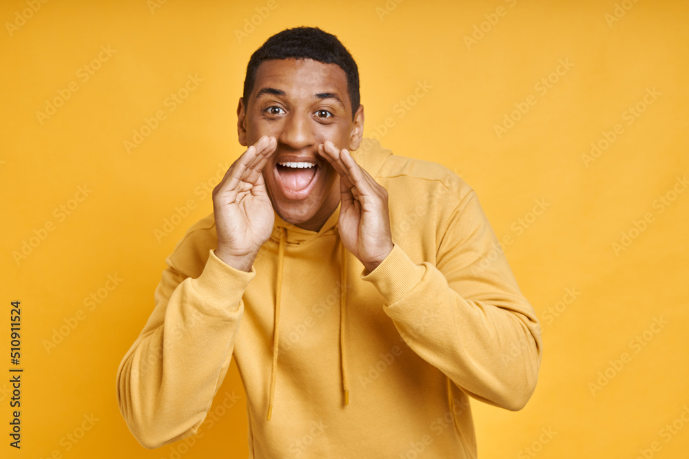Happy young man shouting while covering mouth with hands and standing against yellow background Happy young man shouting while covering mouth with hands and standing against yellow background