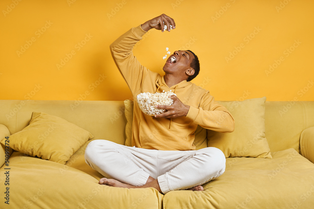 Playful mixed race man enjoying popcorn while relaxing on the couch with yellow background Playful mixed race man enjoying popcorn while relaxing on the couch with yellow background