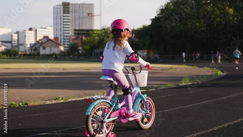 Cute light hair little girl in pink helmet in elbow and knee pads rides a bicycle at the stadium. People run in background. Sport activity for children. Back view photo