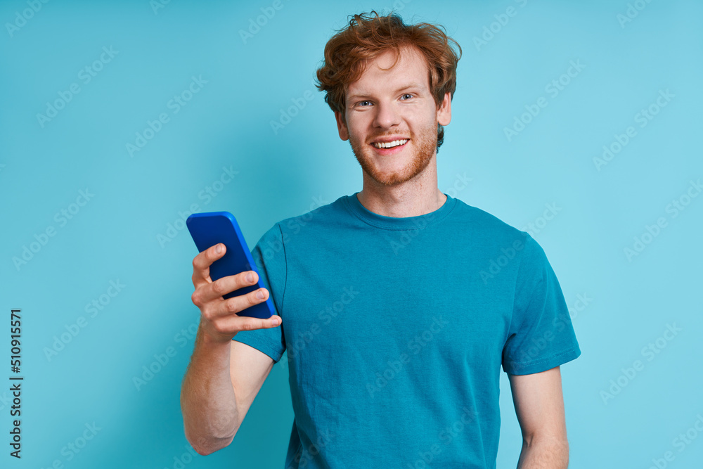 Cheerful redhead man holding mobile phone while standing against blue background Cheerful redhead man holding mobile phone while standing against blue background