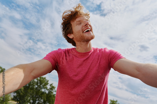 Happy young redhead man making selfie and smiling while standing outdoors