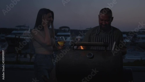 Couple Enjoy Summer Evening at Yacht Club. Man and Woman On Picnic and Barbecue On Beach With Yachts. Man Cooking Grilled Meat On Barbecue Grill. Prepare Food Outdoor On Happy Holidays.