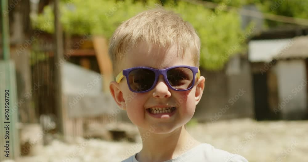 Portrait of a happy child who sincerely smiles at the camera while standing on the street of a country house in sunglasses on his face Portrait of a happy child who sincerely smiles at the camera while standing on the street of a country house in sunglasses on his face