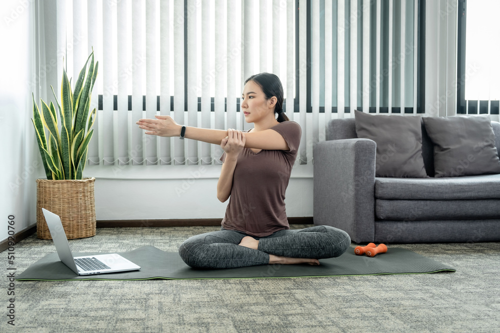 Young woman in sportswear is sitting on mat to stretching arms and body to warm up prepare before yoga exercise in the living room at home Young woman in sportswear is sitting on mat to stretching arms and body to warm up prepare before yoga exercise in the living room at home