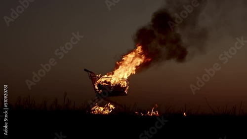 A man pours gasoline from a canister on a burning cart from a supermarket in an empty field, against the background of a golden sunset closer to a dark night, slow motion