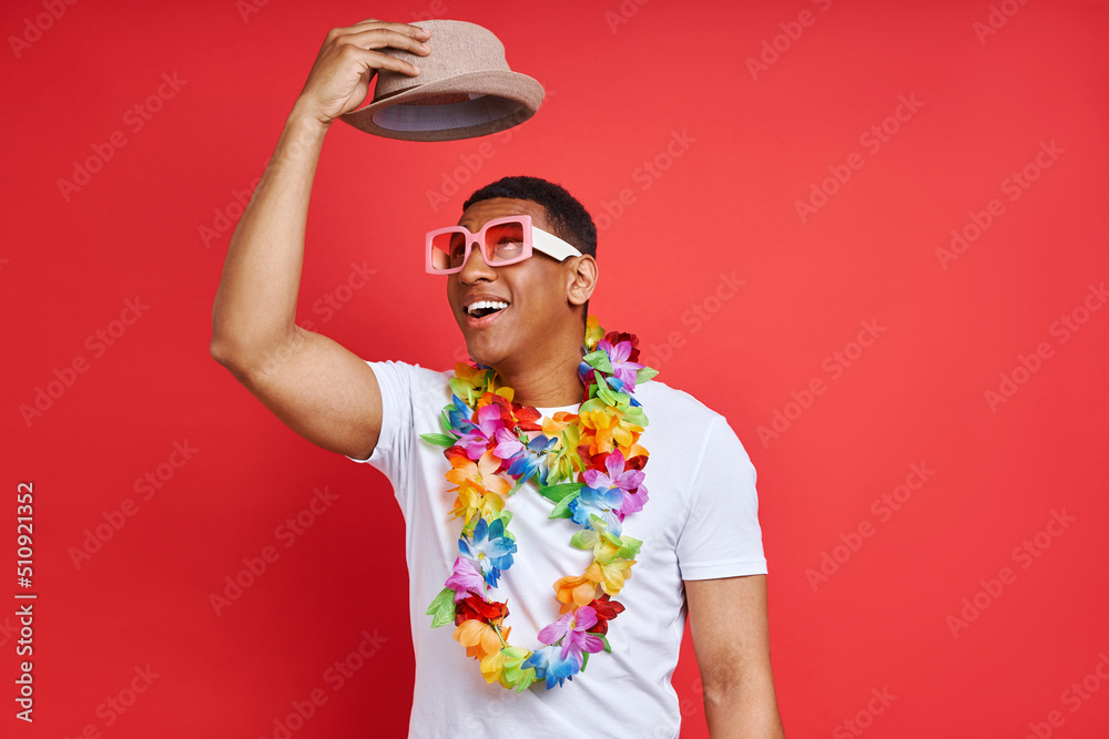 Happy young man in Hawaiian necklace carrying hat while standing against red background Happy young man in Hawaiian necklace carrying hat while standing against red background