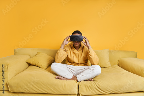 Confident young man in virtual reality glasses relaxing on the couch with yellow background