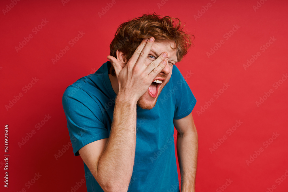 Playful redhead man touching face and grimacing while standing against red background Playful redhead man touching face and grimacing while standing against red background