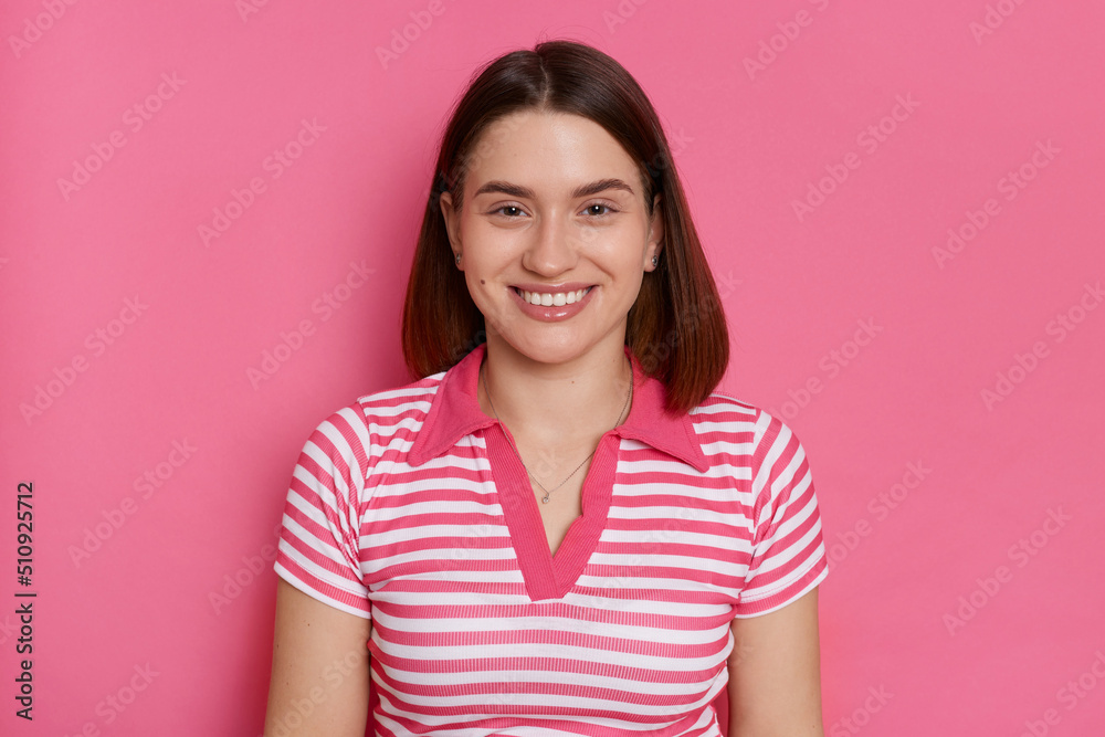 Indoor shot of smiling satisfied woman wearing striped T-shirt looking at camera with toothy smile, expressing positive emotions, posing isolated over pink background. Indoor shot of smiling satisfied woman wearing striped T-shirt looking at camera with toothy smile, expressing positive emotions, posing isolated over pink background.