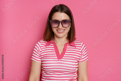 Portrait of delighted happy positive woman wearing striped T-shirt and sunglasses posing isolated over pink background, looking at camera with pleasant facial expression.