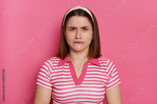 Portrait of scared nervous young Caucasian woman wearing striped T-shirt and hair band posing isolated over pink background, looking at camera and biting lips.