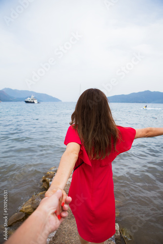 beautiful brunette girl in red dress is standing on the sea and is holding her beloved's hand photo