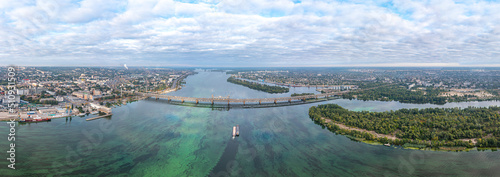 A large laden dry cargo ship is docked on the Dnieper River against the background of a large railway bridge. Large panorama. View from above. Sunny day. Aerial view