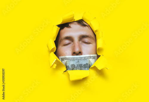 Foto The man's face pokes out through a torn hole in the yellow paper, with a hundred dollar bill and a closed mouth