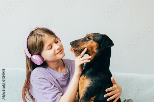 Happy teenage girl in lilac t-shirt sits on the sofa, listens to music has fun playing with the dog