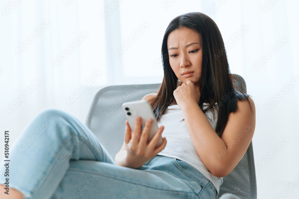 Upset confused tanned lovely young Asian woman read shocked message using phone at home interior living room. Distance communication, Remote work, Social media concept. Cool offer Banner Wide angle Upset confused tanned lovely young Asian woman read shocked message using phone at home interior living room. Distance communication, Remote work, Social media concept. Cool offer Banner Wide angle