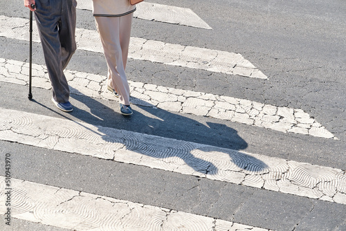 two unrecognizable elderly people, one of them with a cane, crossing a pedestrian crossing