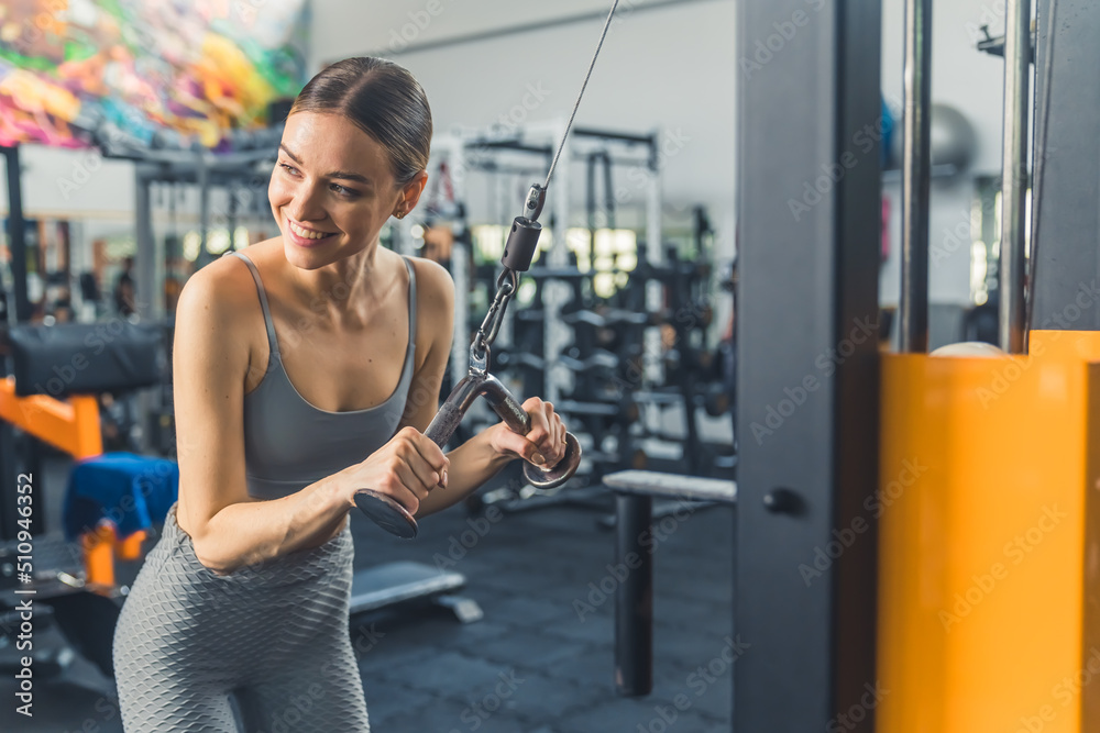 Fototapeta premium Indoor gym shot of a skinny caucasian young adult woman with slicked back hair trying out professional gym equipment for the first time. Arms exercises. High quality photo