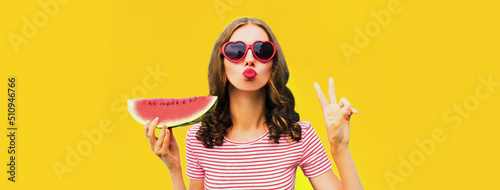 Summer portrait of beautiful young woman blowing her lips with red lipstick sending air kiss with slice of watermelon wearing heart shaped sunglasses on yellow background